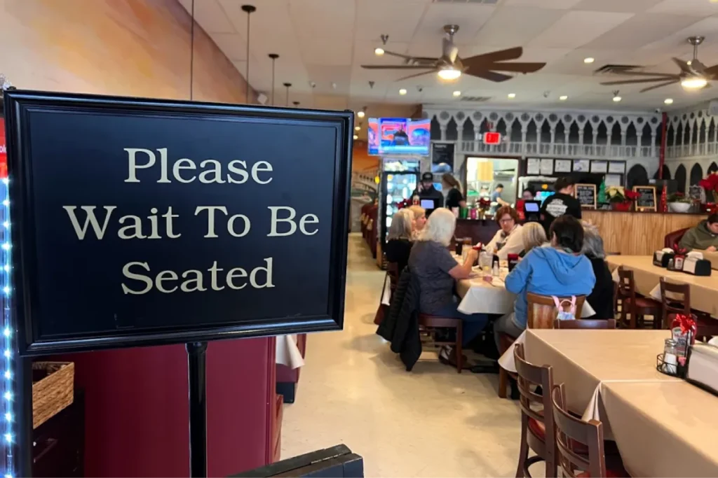 "Please Wait To Be Seated" sign in a restaurant with customers at tables in the background.